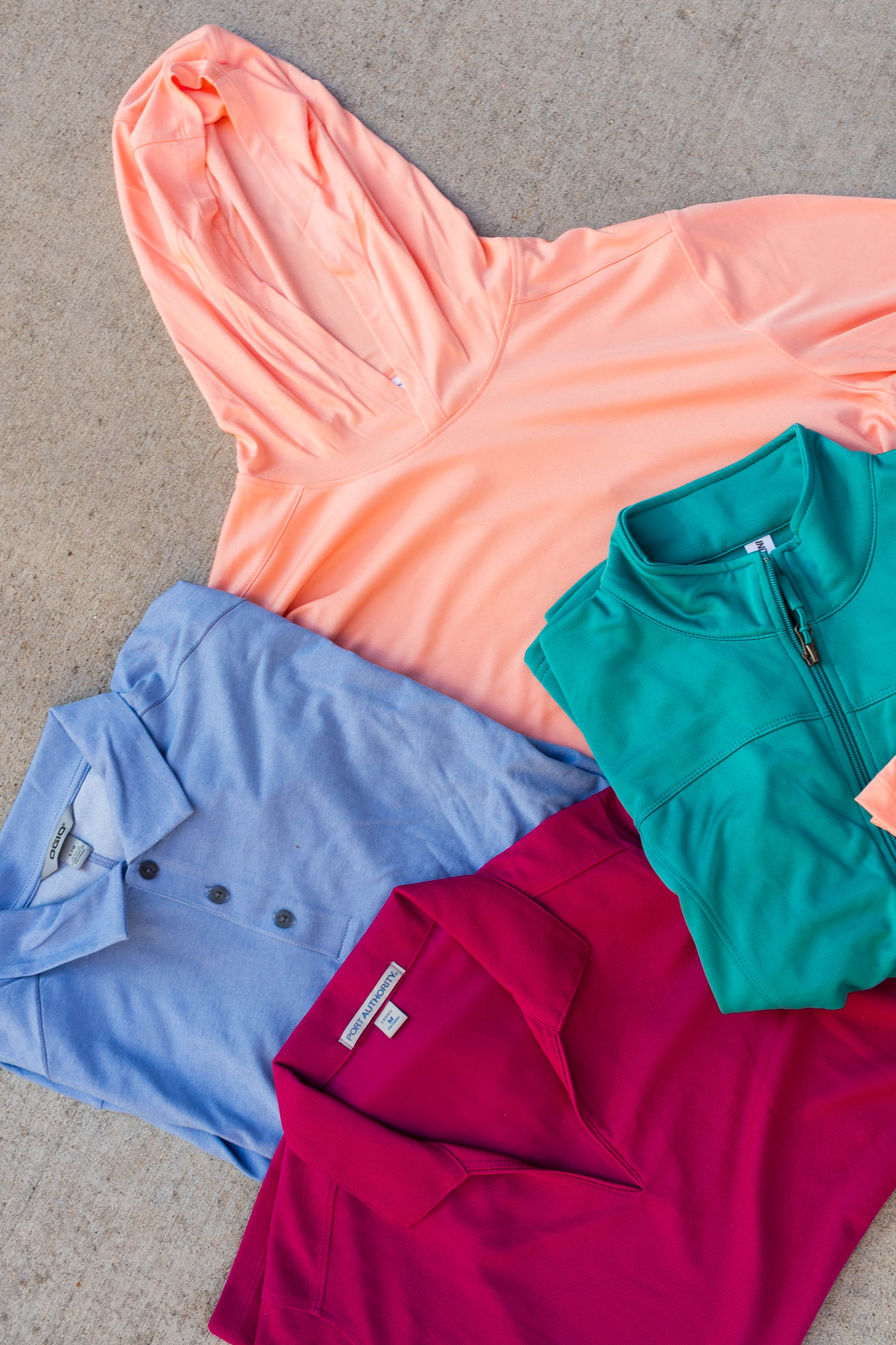 Four folded clothing items arranged on a concrete surface, including a peach-colored hooded sweatshirt, a teal quarter-zip pullover, a light blue buttoned polo, and a deep red collared polo.