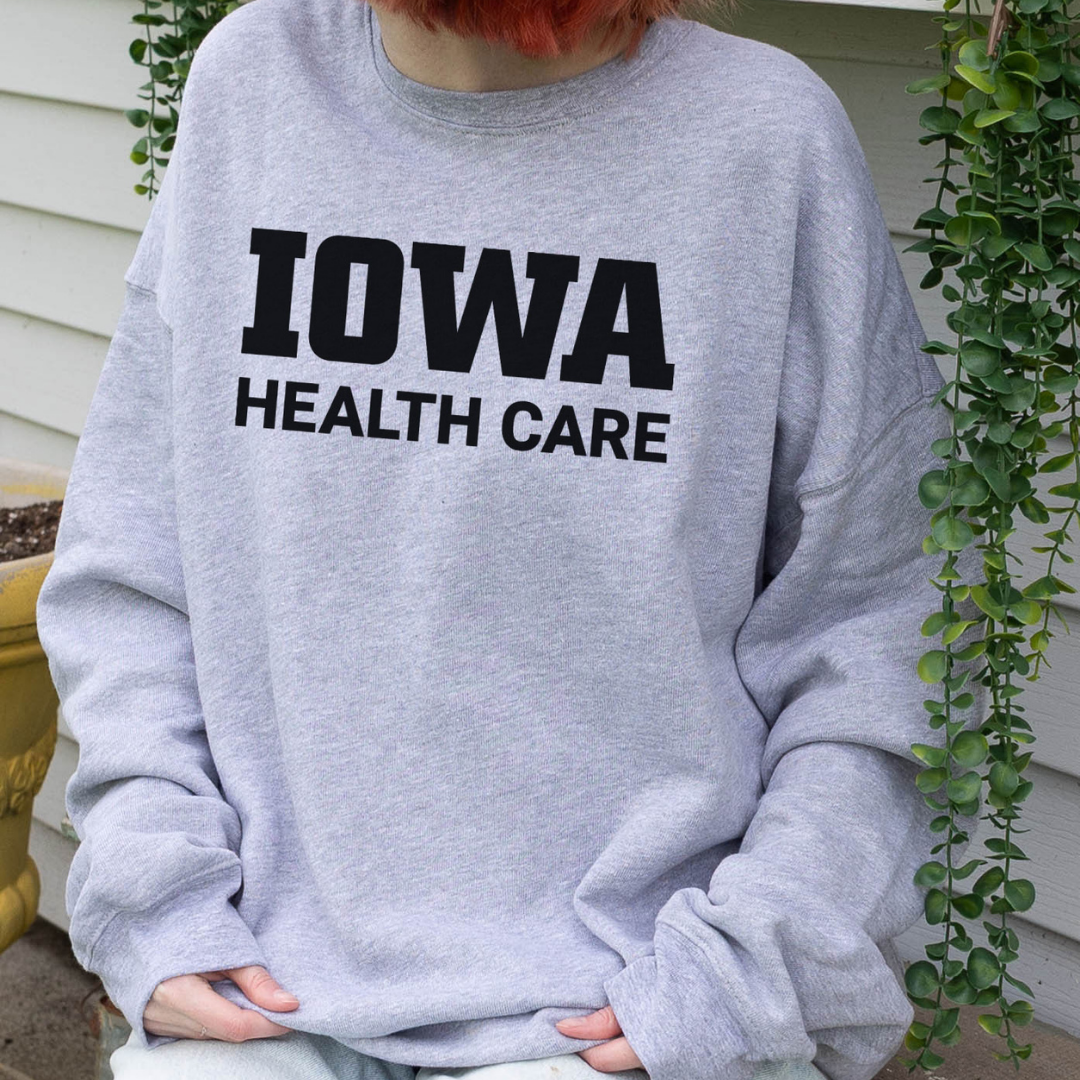 Person wearing a light gray crewneck sweatshirt with bold black text on the front that reads ‘Iowa Health Care,’ seated outside near a house with hanging greenery.