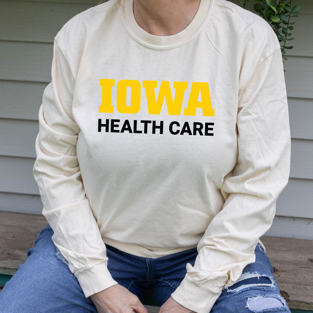 Person wearing an ivory long-sleeve t-shirt with bold yellow and black text on the front that reads ‘Iowa Health Care,’ seated outdoors in ripped blue jeans.