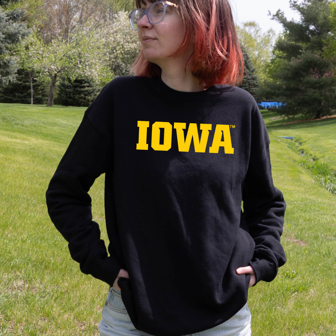 Person standing outdoors on green grass wearing a black crewneck sweatshirt with bold yellow text that reads ‘Iowa.