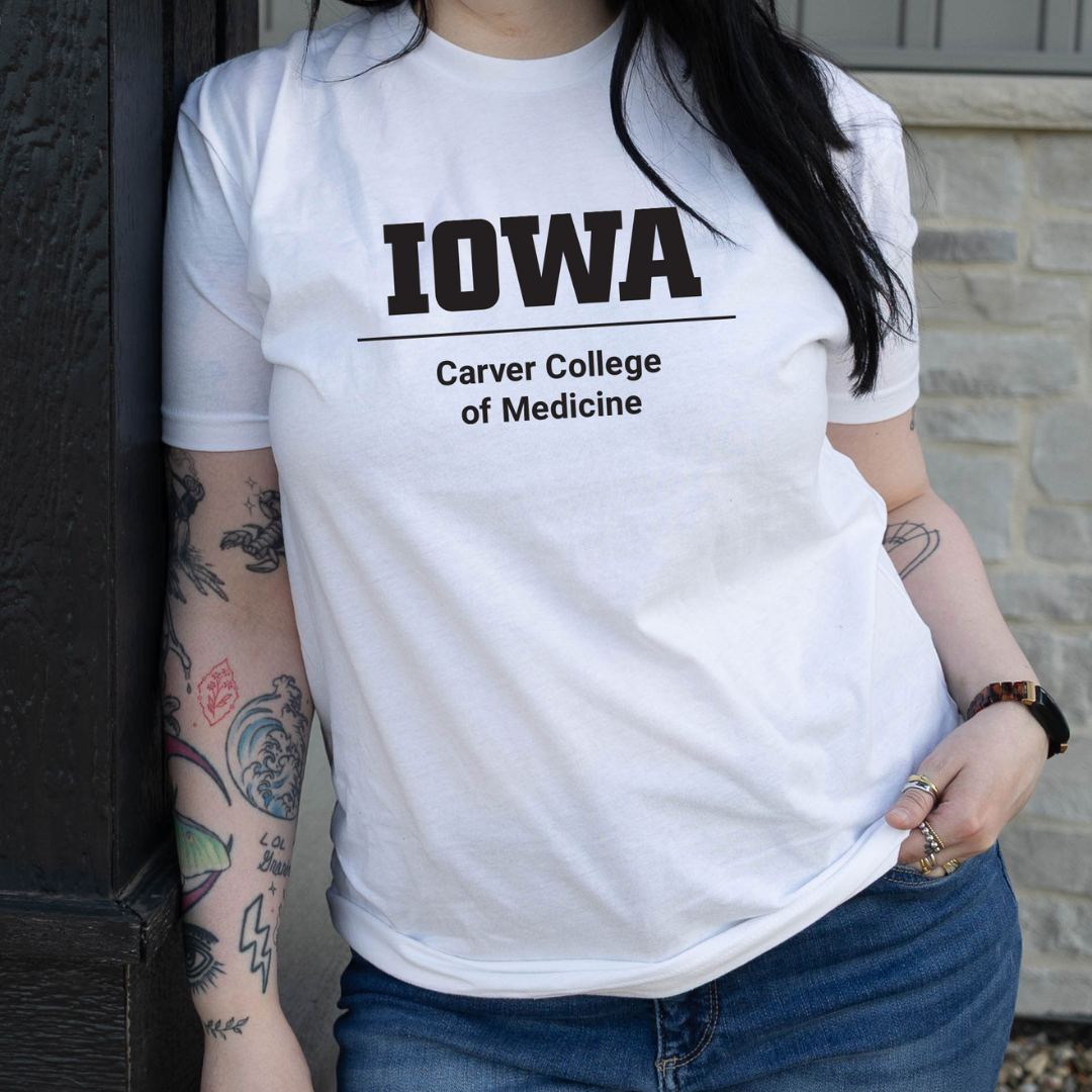 Person wearing a white t-shirt with bold black text that reads ‘Iowa Pharmacy,’ leaning casually against a wall with visible tattoos on their arm.
