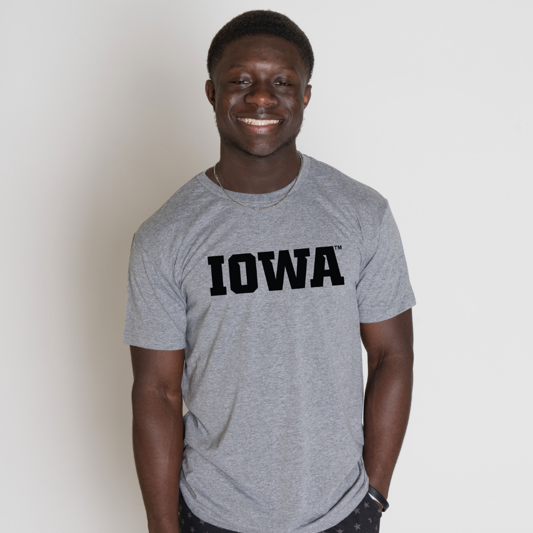 Smiling student wearing a gray t-shirt with bold black text that reads ‘IOWA,’ standing against a plain light background.