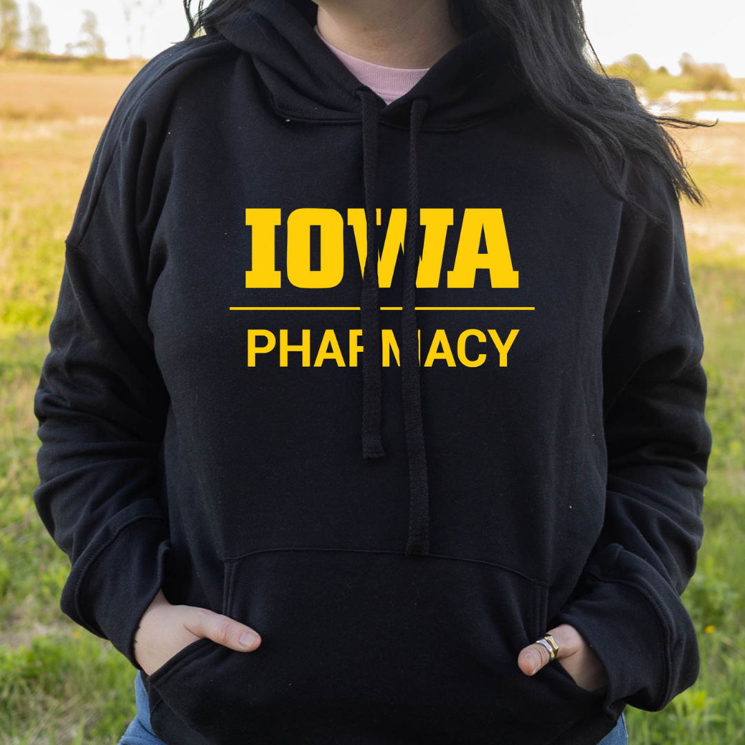 Person wearing a black hoodie with bold yellow text that reads ‘Iowa Pharmacy,’ standing outdoors in a grassy field.