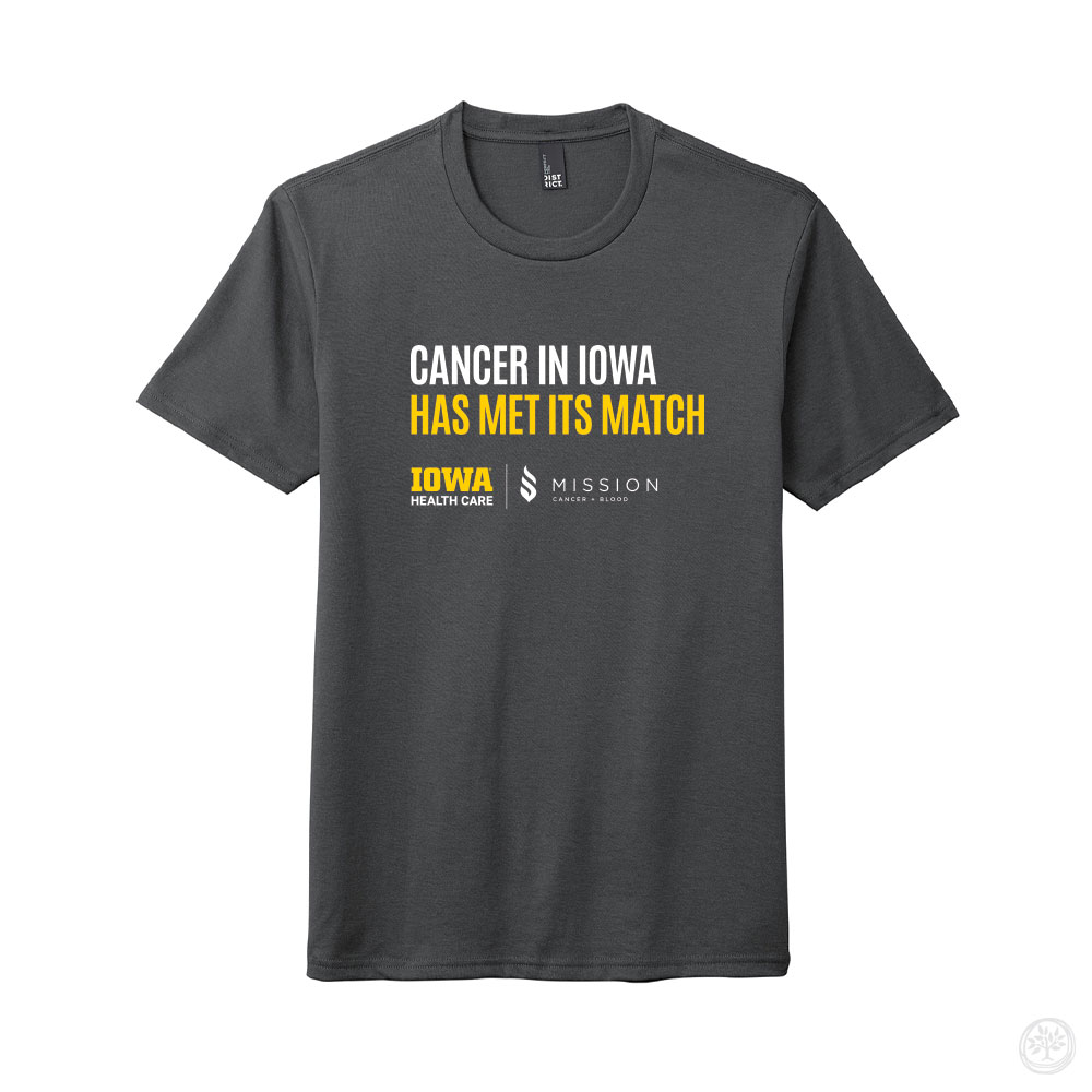 Dark gray t-shirt with bold white and yellow text that reads ‘Cancer in Iowa Has Met Its Match,’ featuring Iowa Health Care and Mission Cancer + Blood logos beneath.