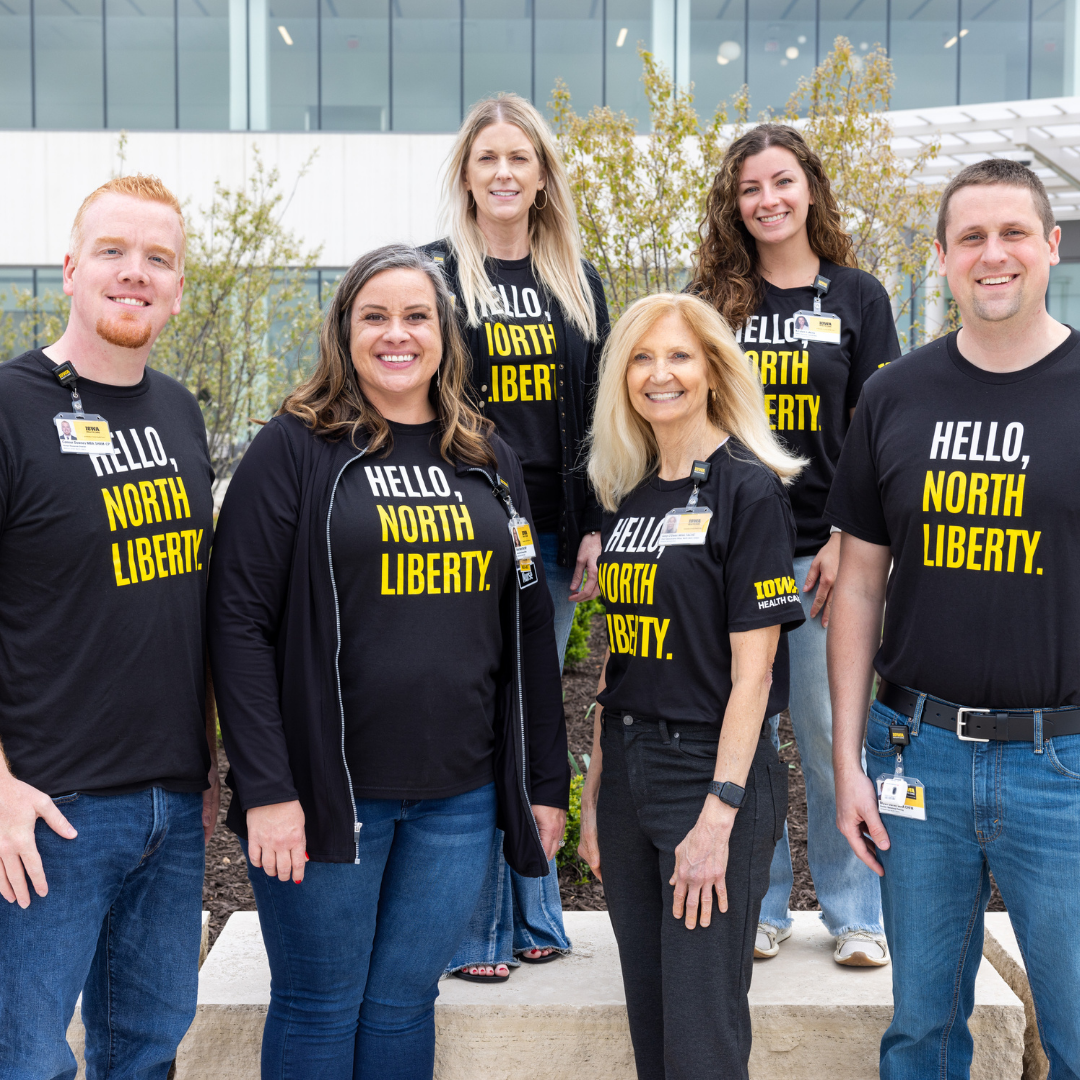 Group of University of Iowa hospital staff smiling outside the main entrance, wearing matching black custom t-shirts with bold yellow text reading “Hello, North Liberty.” Bulk order apparel created for hospital staff unity and community outreach.