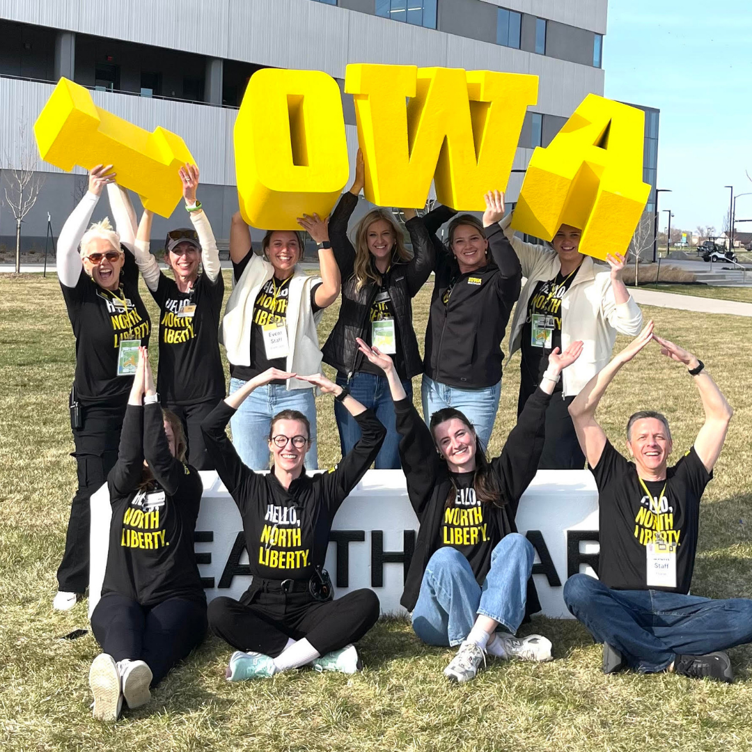 Group of smiling volunteers outside the University of Iowa Hospitals and Clinics holding large yellow block letters spelling 'IOWA,' wearing matching black 'Hello North Liberty' t-shirts for a bulk hospital project.