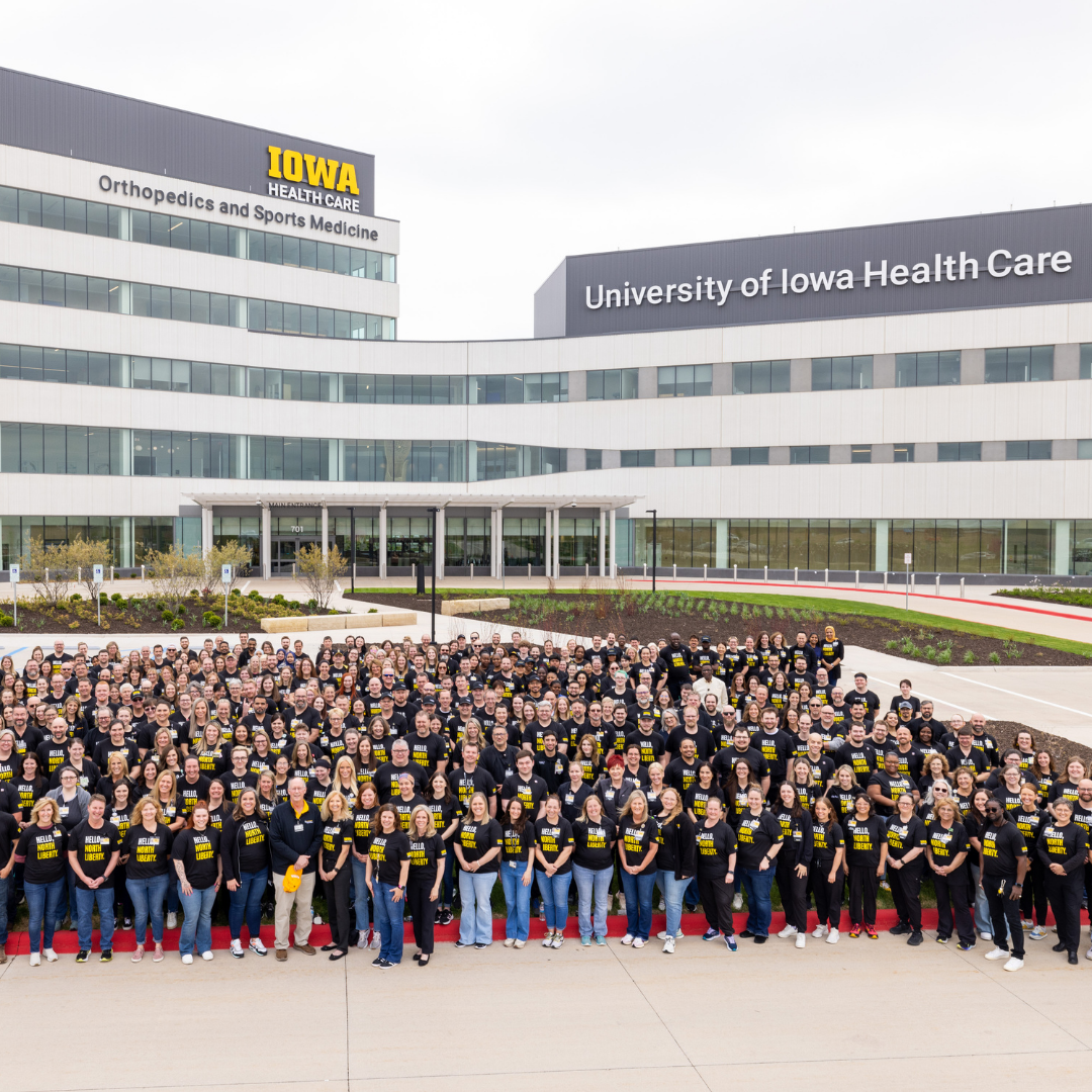 Large group of University of Iowa Health Care staff standing together outside the Orthopedics and Sports Medicine building, all wearing matching black custom t-shirts with bold yellow text reading “Hello, North Liberty.” Bulk apparel order showcasing hosp