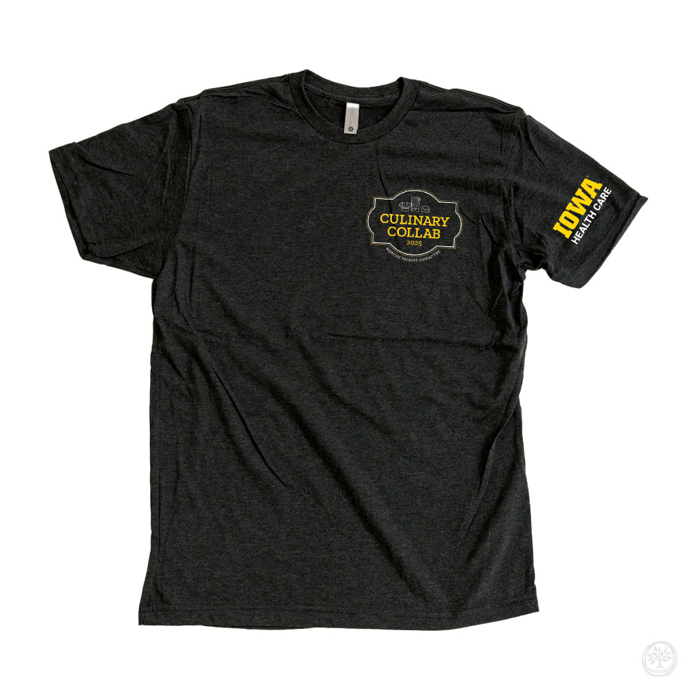 Black t-shirt with yellow text reading ‘Culinary Collab’ and white text below saying ‘Marcom Retreat Committee,’ featuring chef hat graphics above. The Iowa Health Care logo is printed in yellow on the sleeve.